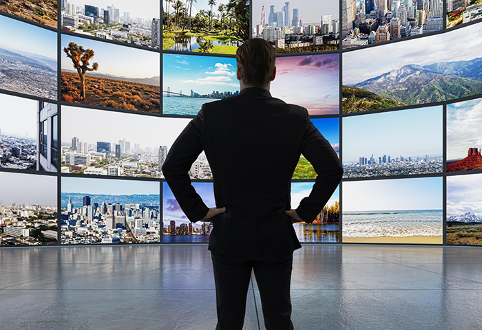 Businessman viewing multiple city landscapes on large digital screens in modern control room